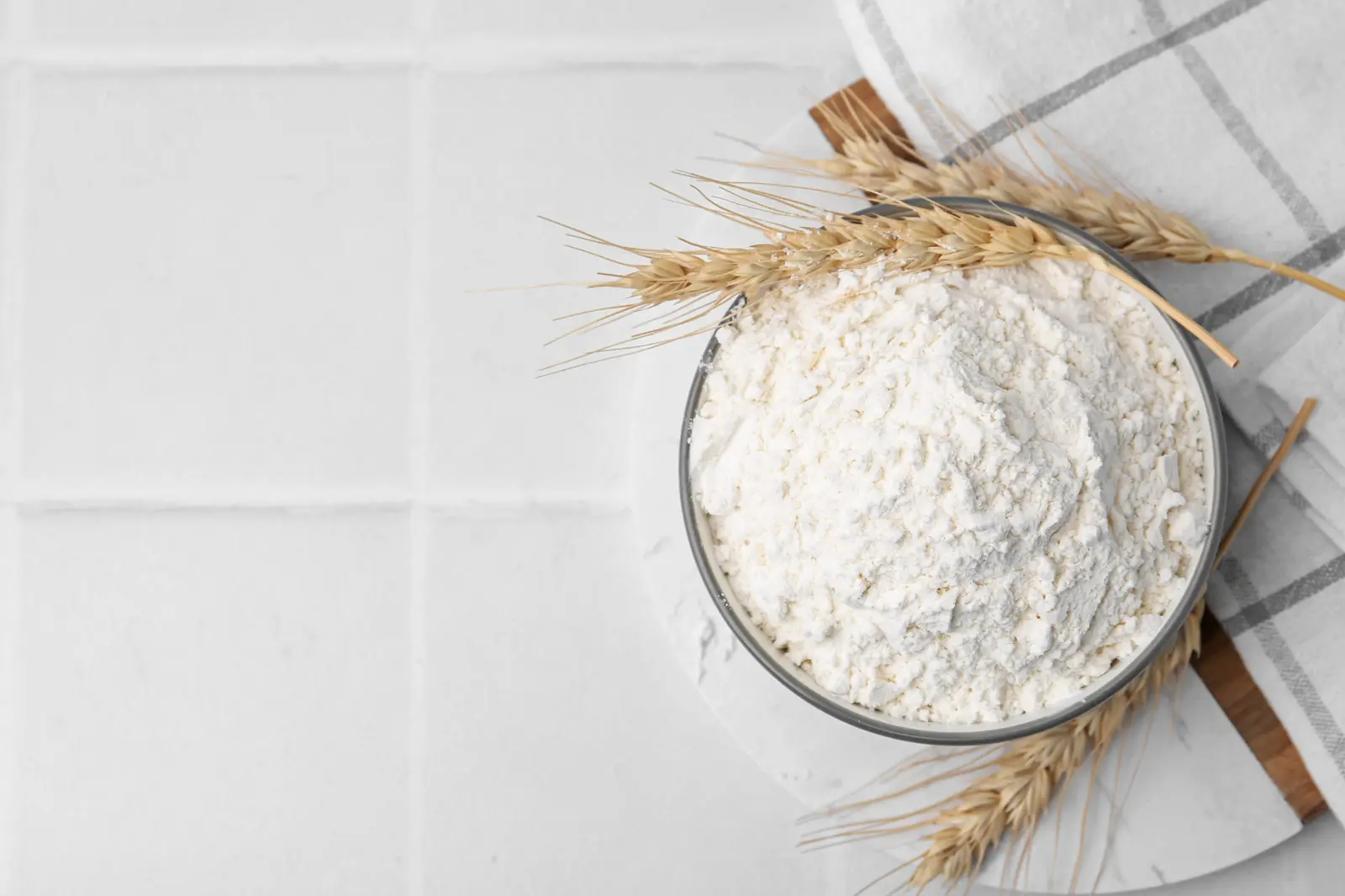 Bowl of fresh wheat flour with wheat sprigs on a white tile surface