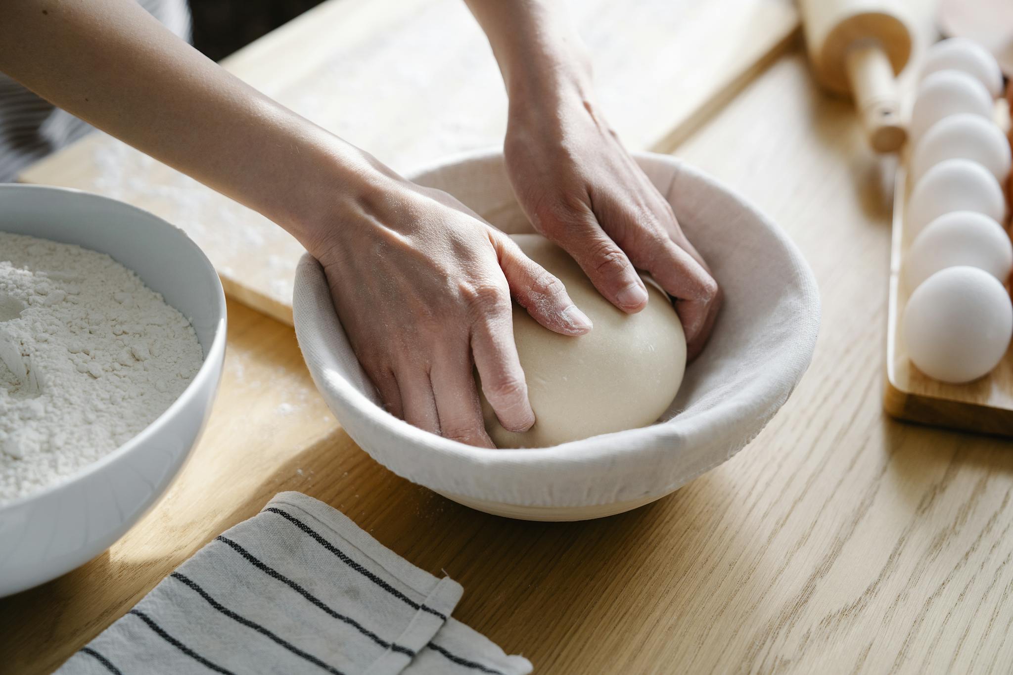 Hands kneading dough on a wooden table with flour and eggs, capturing a cozy baking scene.